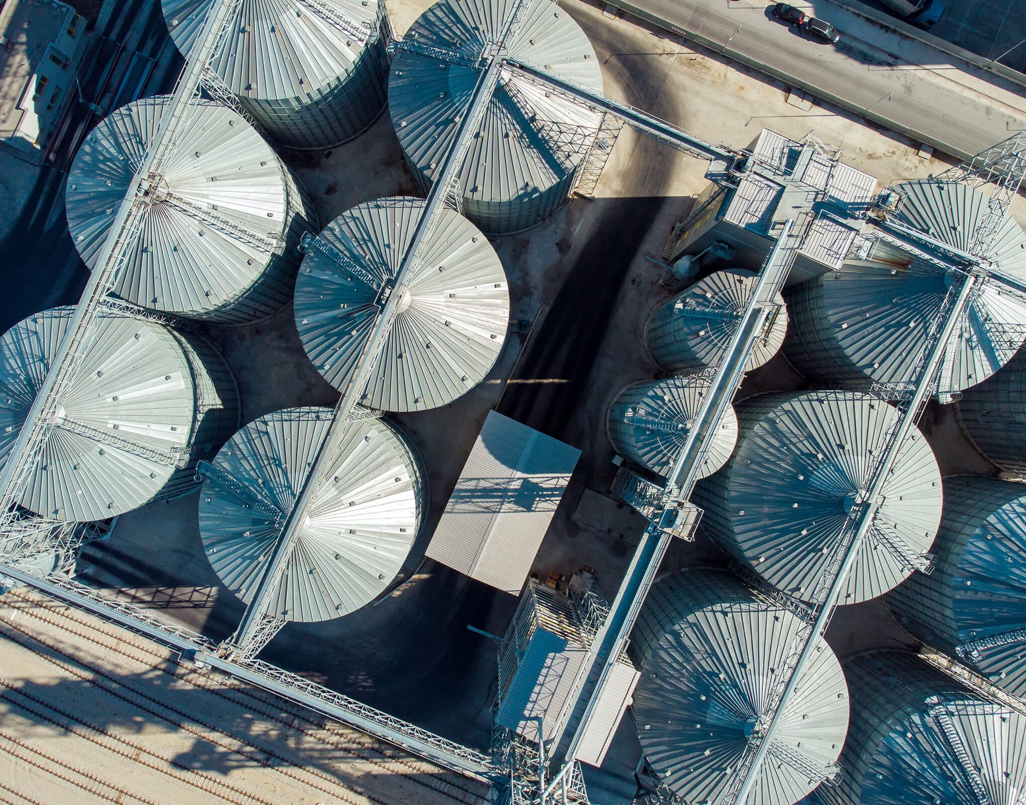 Aerial View of Grain Silos in Industrial Area Aerial perspective of large metallic grain silos placed in a row, showcasing their circular design and surrounding infrastructure.