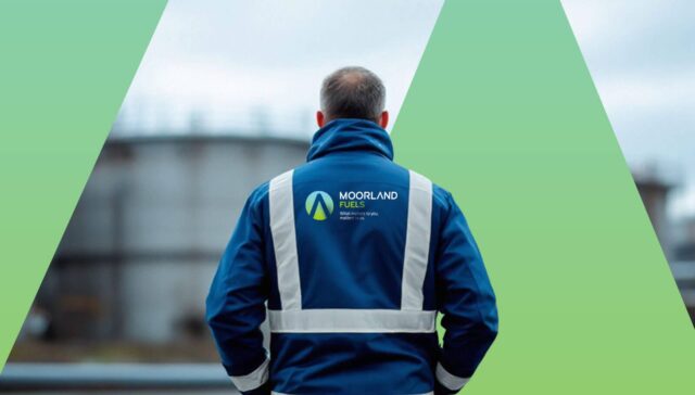 A man wearing a blue safety jacket with reflective straps, standing with his back to the camera, looking at an industrial landscape.