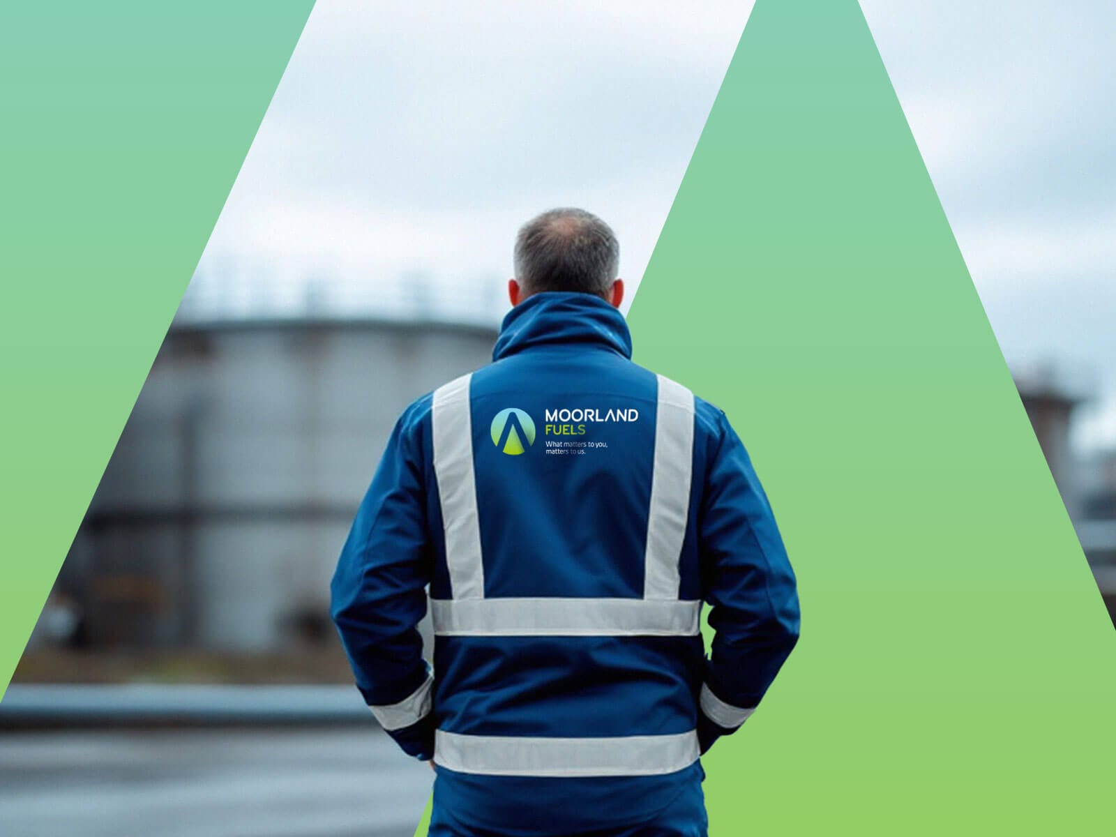 A man wearing a blue safety jacket with reflective straps, standing with his back to the camera, looking at an industrial landscape.