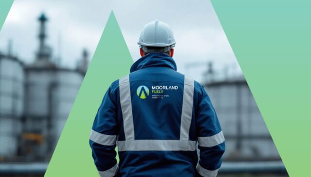 A worker wearing safety attire and a hard hat, standing with their back to the camera at an industrial site with storage tanks in the background.