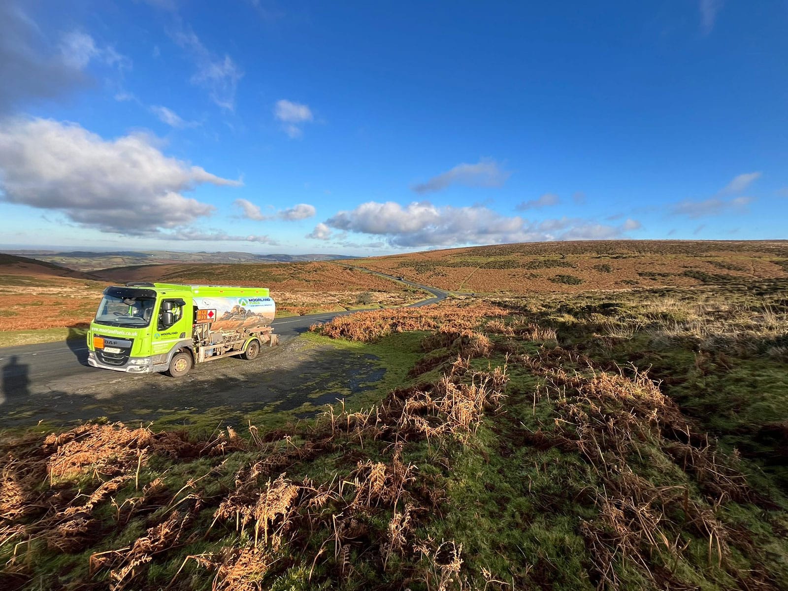 A delivery van parked on a grassy road surrounded by rolling hills and blue sky with scattered clouds.